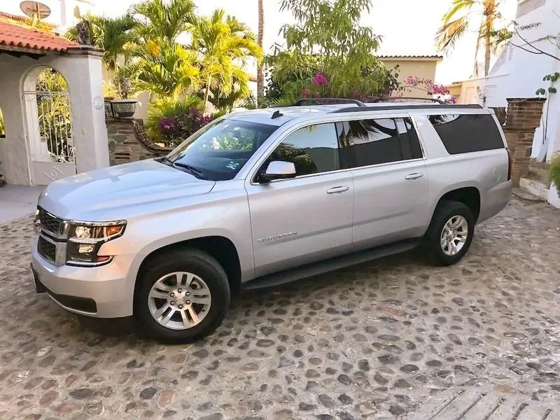 Silver Chevrolet Suburban parked on a cobblestone driveway with a white wall, archway, and tropical plants in the background.
