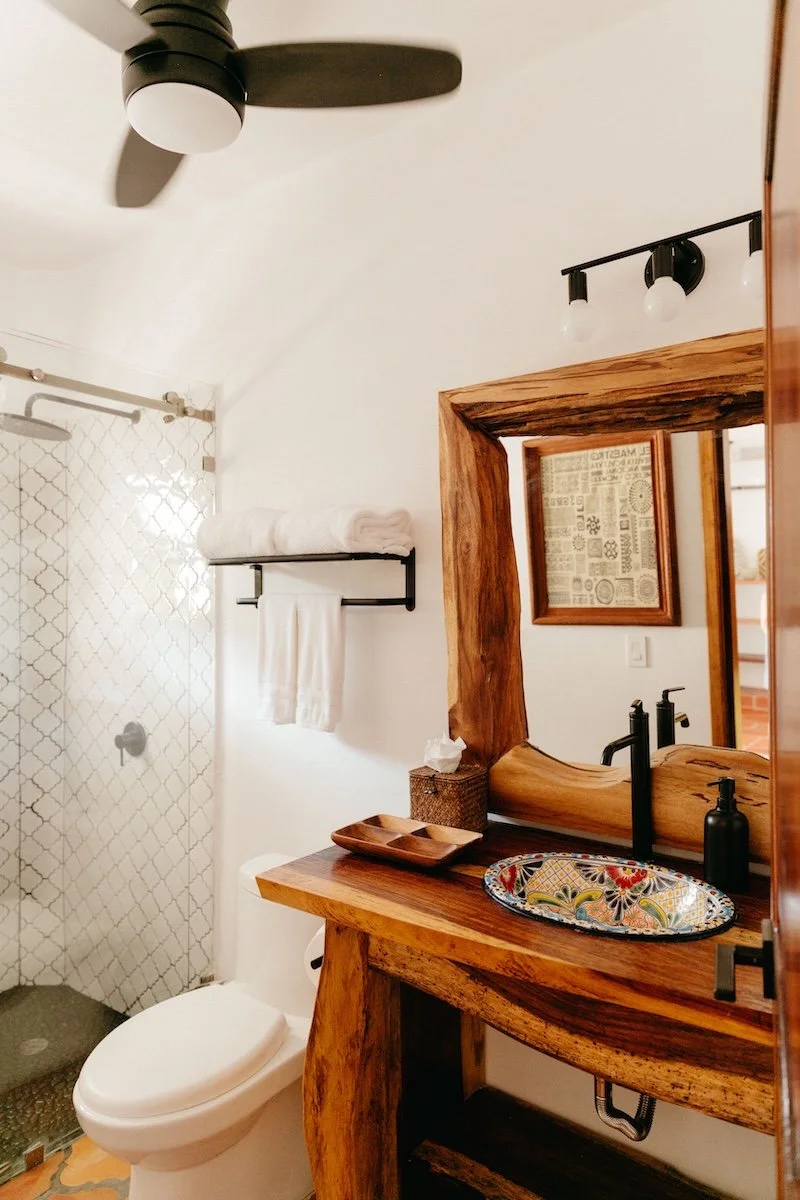 A bathroom with a wooden vanity, colorful sink, black faucet, large mirror, towel rack, toilet, and a shower with a glass door and white patterned tiles.