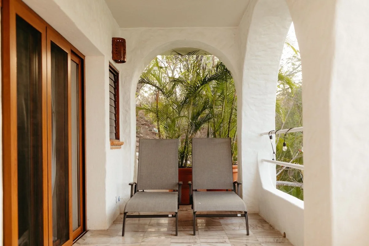 Two patio chairs on a balcony with potted plants and trees in the background.