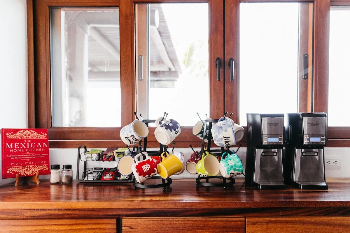 Kitchen countertop with two coffee makers, a mug holder with colorful mugs, a red cookbook titled 'The Mexican Home Kitchen' by Mely Martinez, and two small spice jars. There's a window behind the countertop.