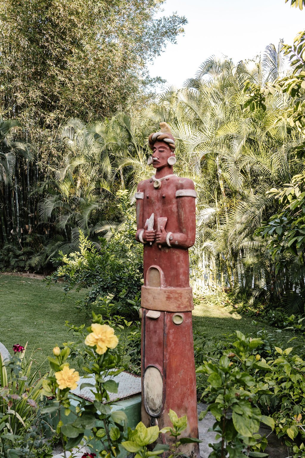 A colorful tribal-style sculpture of a person with elaborate headgear and large earrings, standing in a lush garden with tropical plants and flowers.
