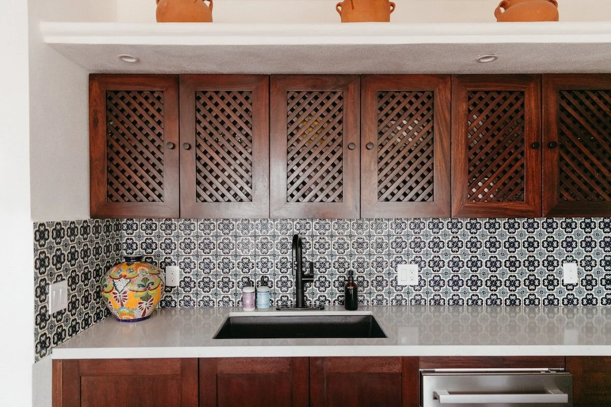 Kitchen with dark wood cabinets, decorative tile backsplash, black sink, gold and purple soap dispensers, and a colorful vase on the counter.