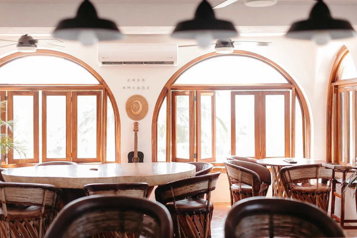 Cozy dining area with wooden tables and wicker chairs, sunlit from large arched windows, wall decor featuring a straw hat, a guitar, and the words 'Casa Amigos', with an air conditioning unit above.