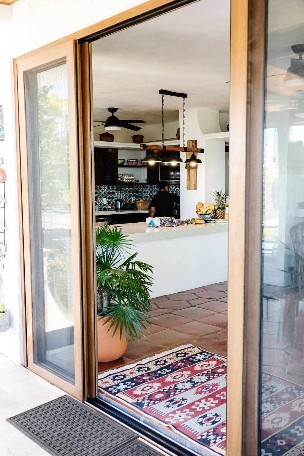 View through a sliding glass door into a kitchen with a colorful patterned rug, a potted plant, a white kitchen counter, and a person working in the kitchen.