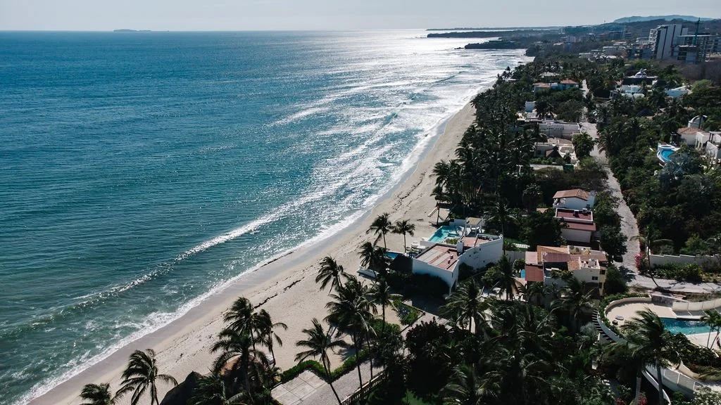 An aerial view of a beach with clear blue water, white sand, and a row of houses and palm trees lining the shore.