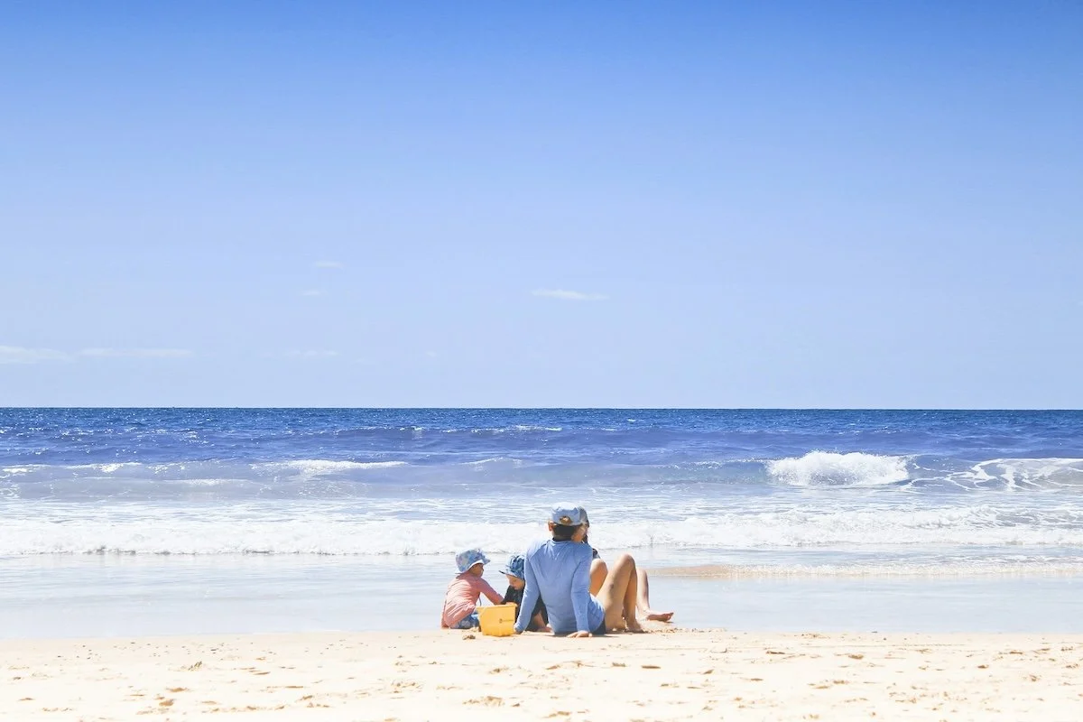 Woman with two children sitting on a sandy beach near the ocean, under a clear blue sky.