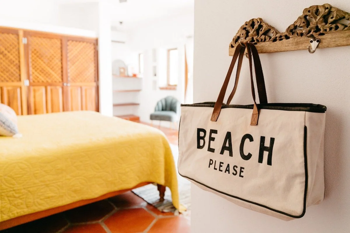 A beige tote bag with black lettering that reads "BEACH PLEASE" hangs from a decorative wooden wall hook in a bedroom with a yellow quilted bedspread.