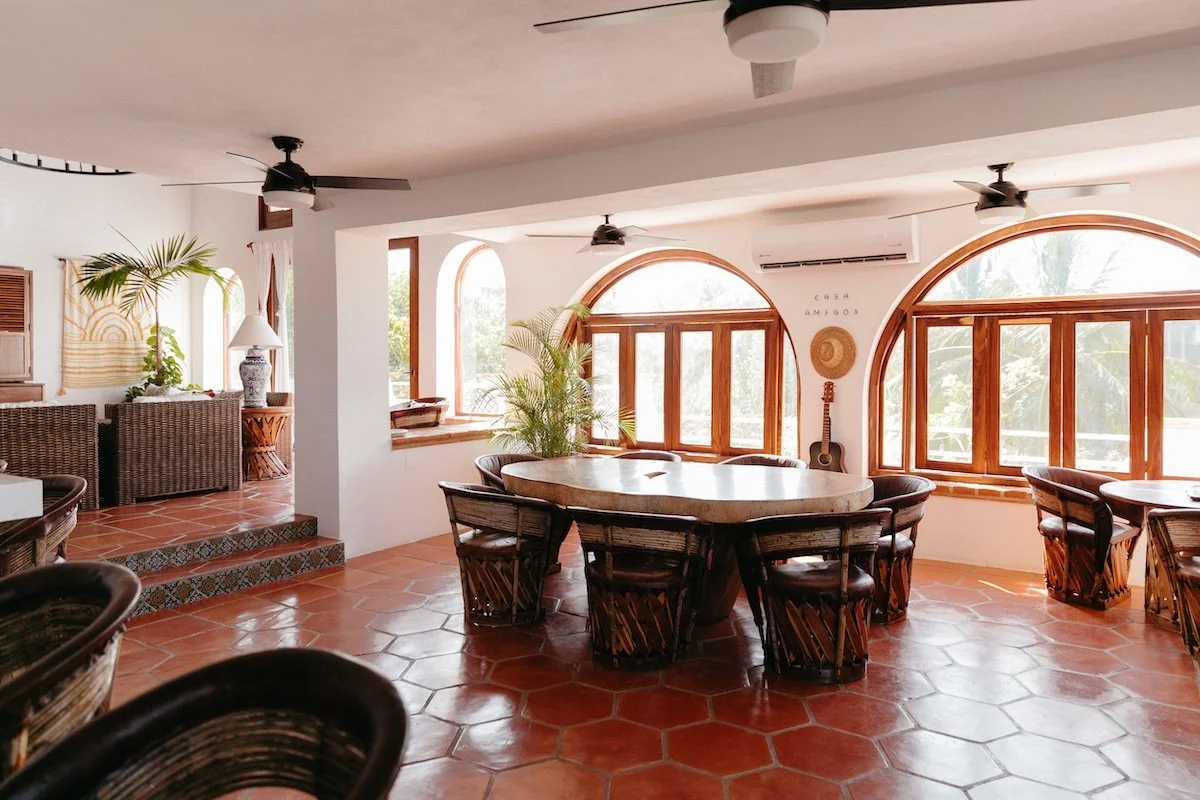 Interior of a bright, tropical-style living room with large arched windows, wicker furniture, green plants, and wooden accents.