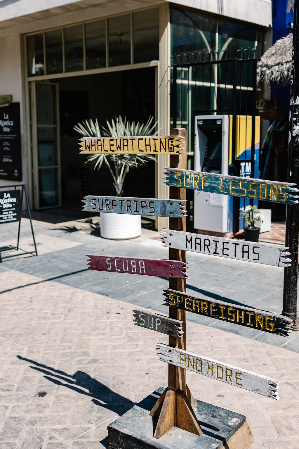 Colorful wooden signs with words like 'WHILE WAITING,' 'SURF LESSONS,' 'SURF TRIPS,' 'MARLIETAS,' 'SCUBA,' 'SPEARFISHING,' 'SUP,' and 'AND MORE,' standing on a sidewalk outside a building.