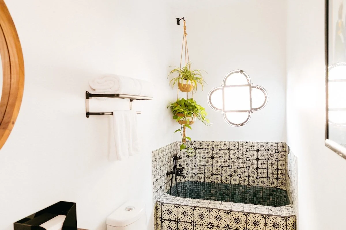 Bathroom with a black tiled bathtub, hanging plants, a window with a geometric frame, a shelf with towels, and a toilet.