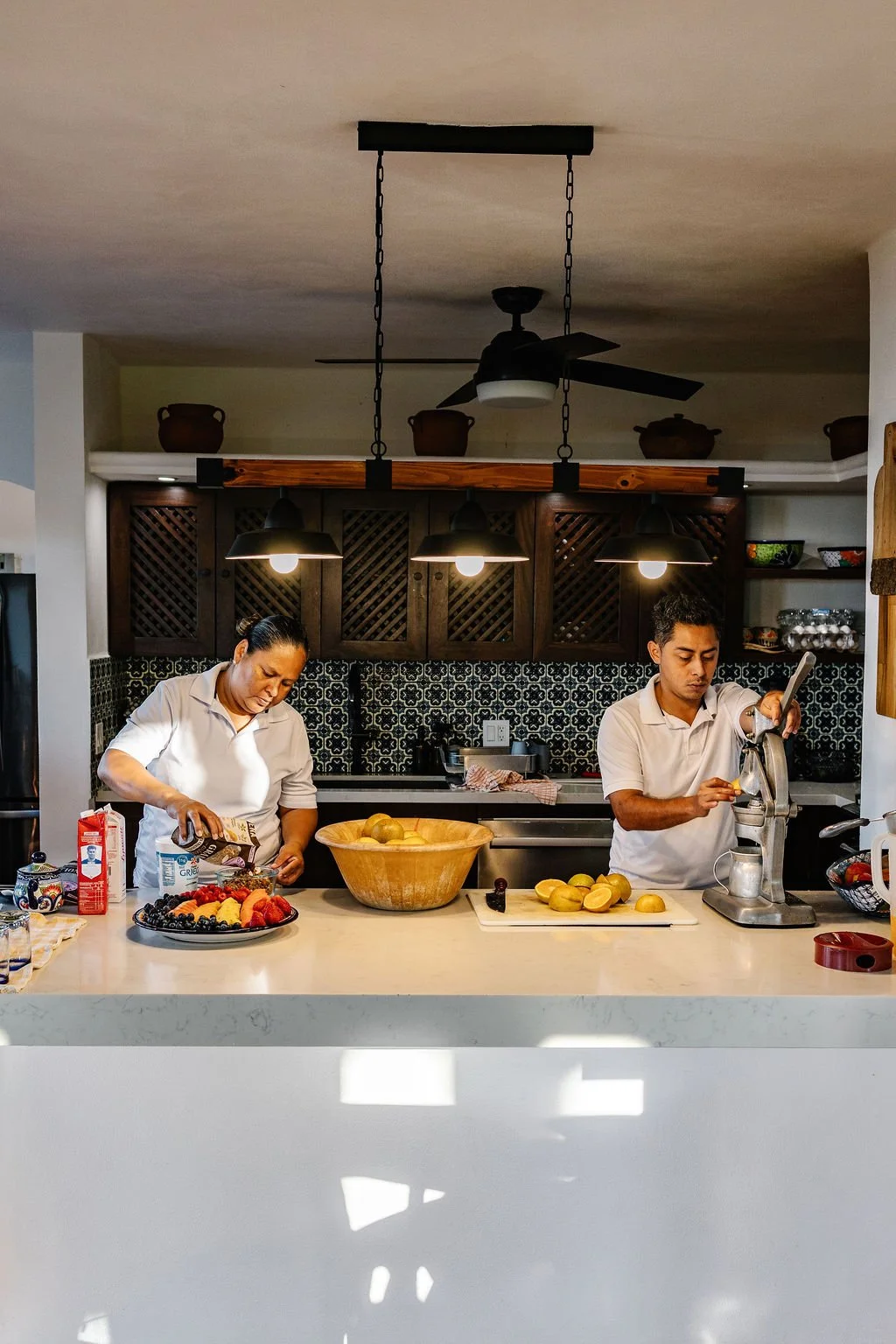 Two people preparing fruit in a kitchen, with a large bowl of assorted fruit and a fruit slicer on the countertop.