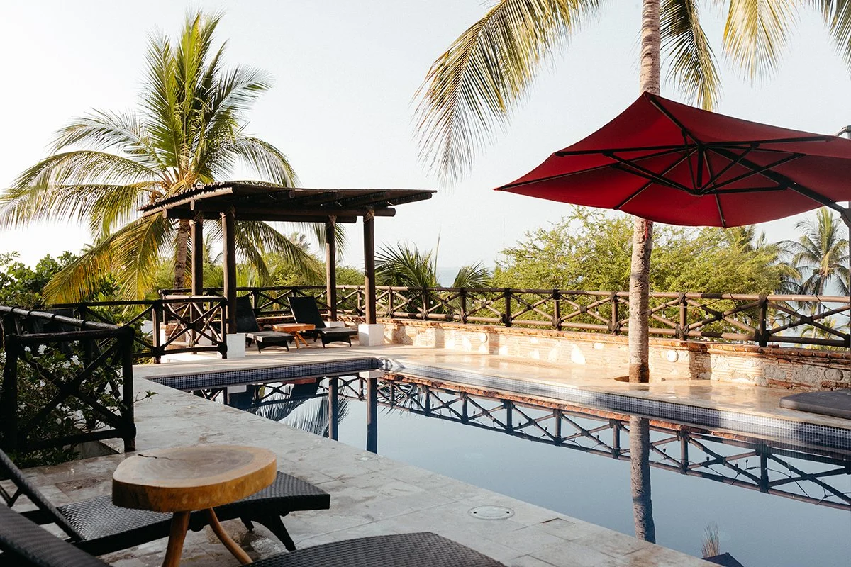 A tropical poolside scene with a small pool reflecting the sky, surrounded by lounge chairs, an umbrella, palm trees, and a wooden gazebo.