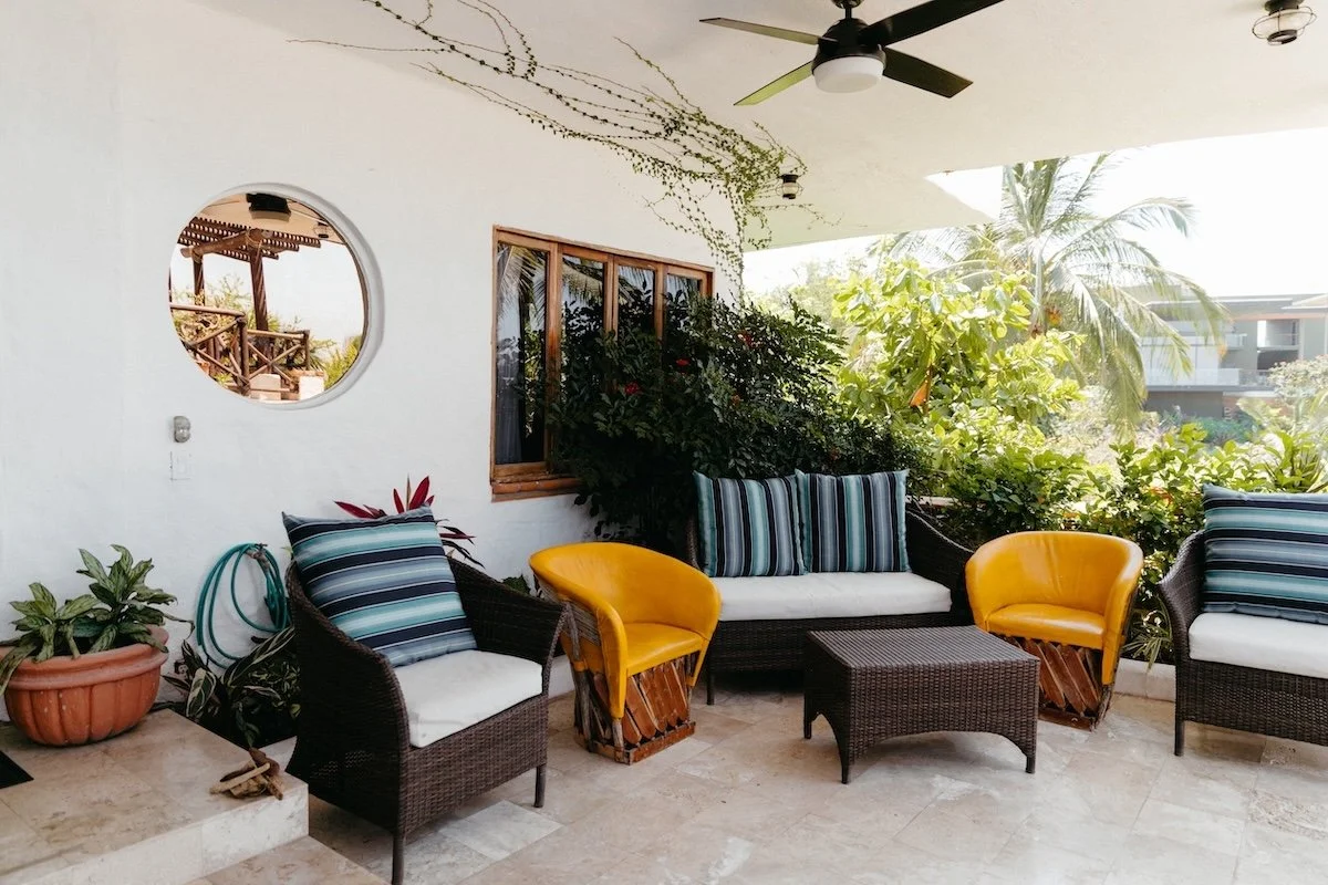 Outdoor patio with wicker furniture including lounge chairs and a sofa, yellow and striped cushions, potted plants, greenery, a ceiling fan, and a round mirror on a white wall.