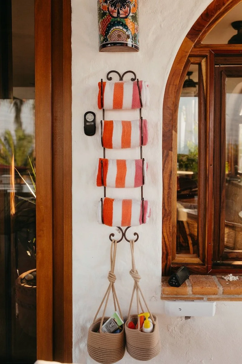 Wall-mounted organizer with four striped towels in orange, pink, and white. A decoration with a butterfly design is above, and two beige baskets with items hang below. A window with wooden trim is on the right.