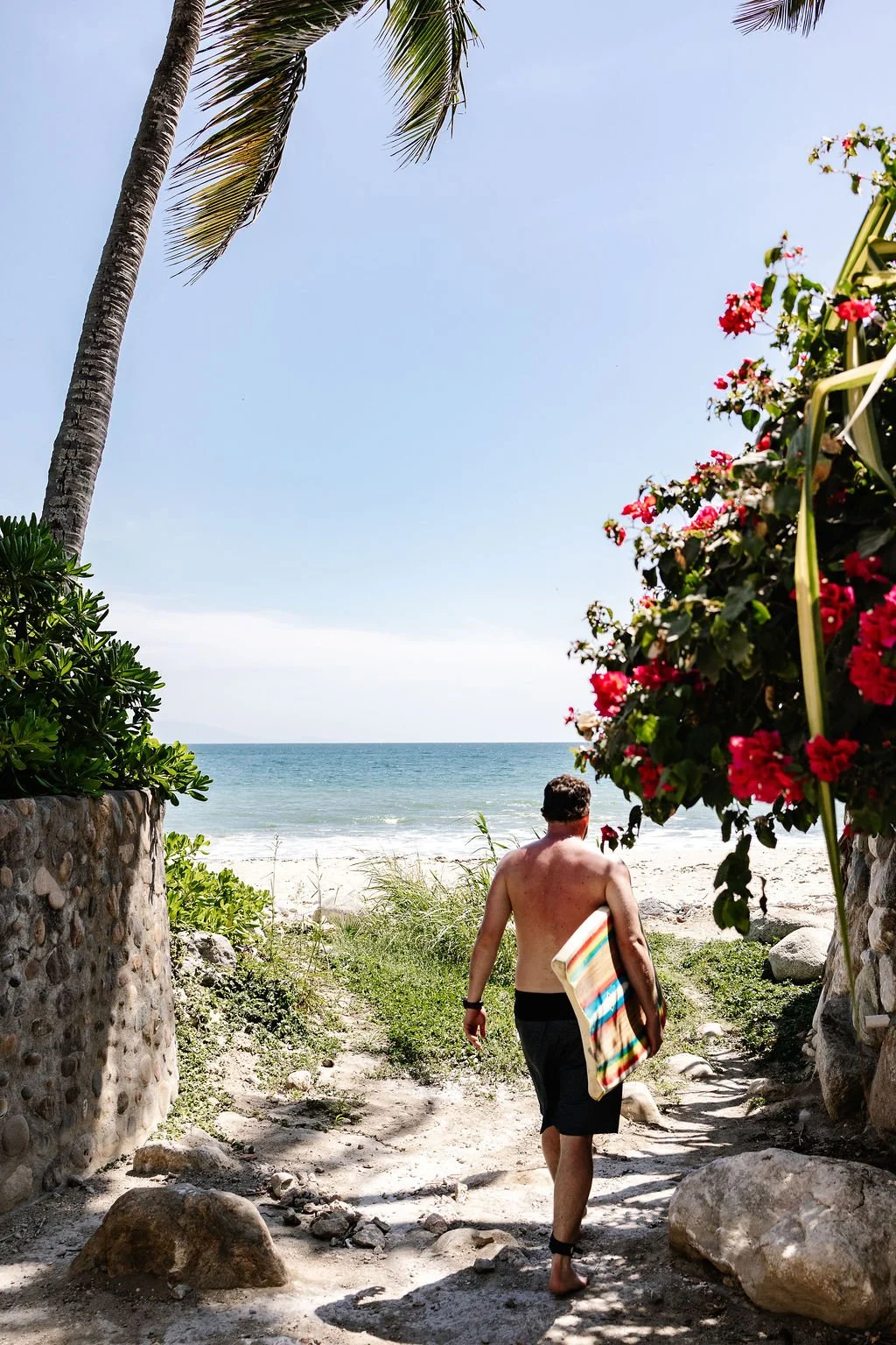 A couple holding hands walking along a sandy beach near the ocean, with palm trees and houses in the background under a clear blue sky.