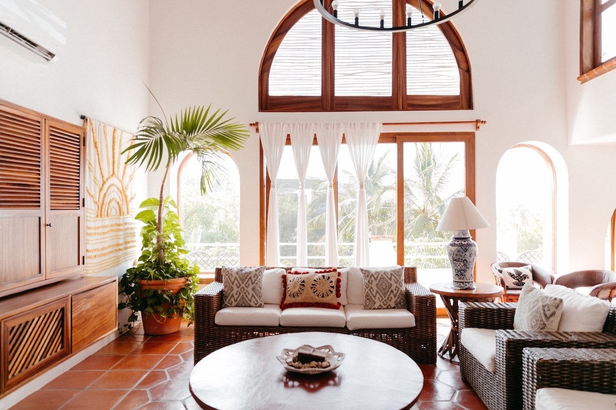 Bright living room with large windows, white curtains, wicker furniture with cushions, a round coffee table, a table lamp, and tropical plants.
