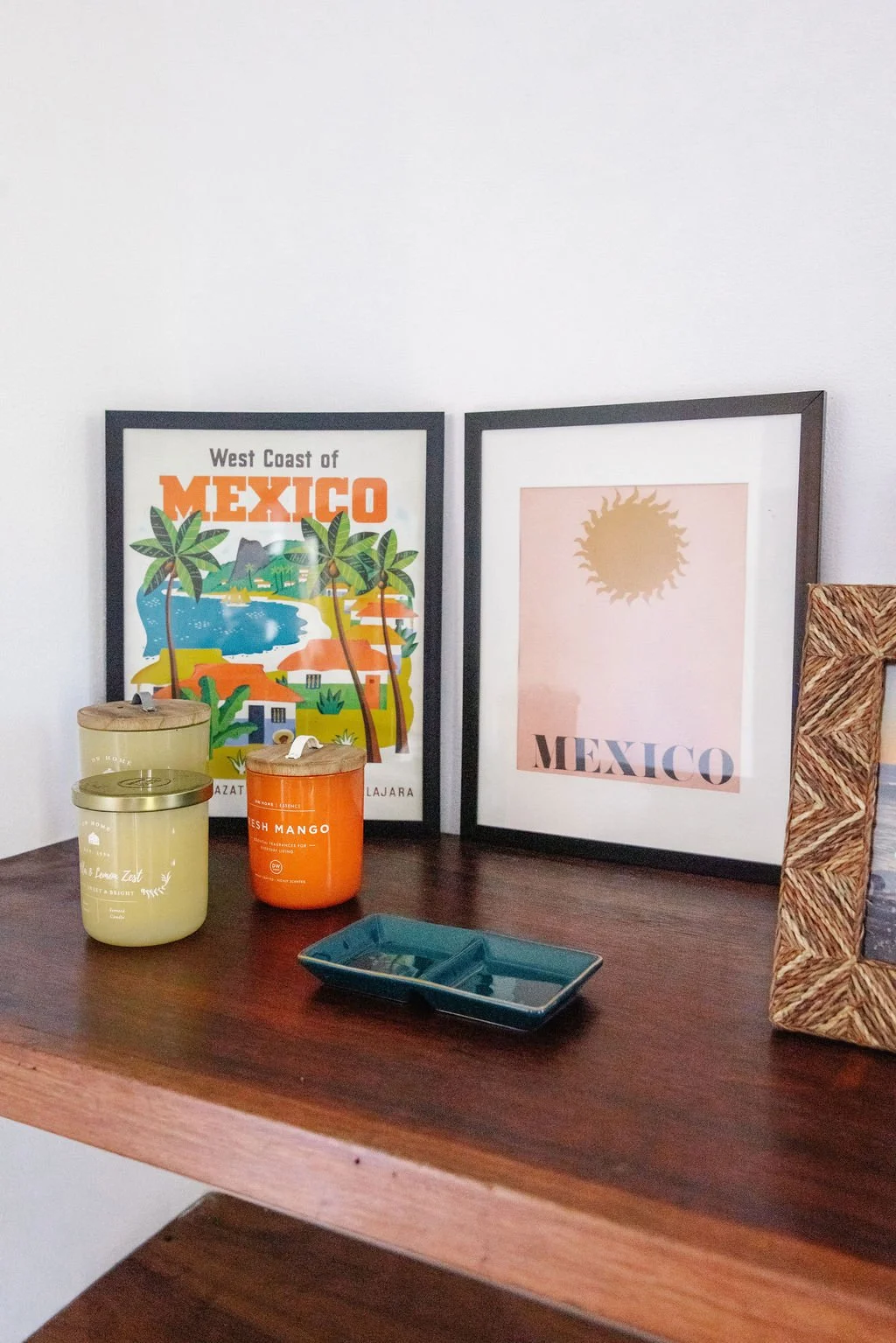 Decorative items on a wooden surface, including framed artwork with a Mexico theme, candles, a small blue dish, and a woven picture frame.