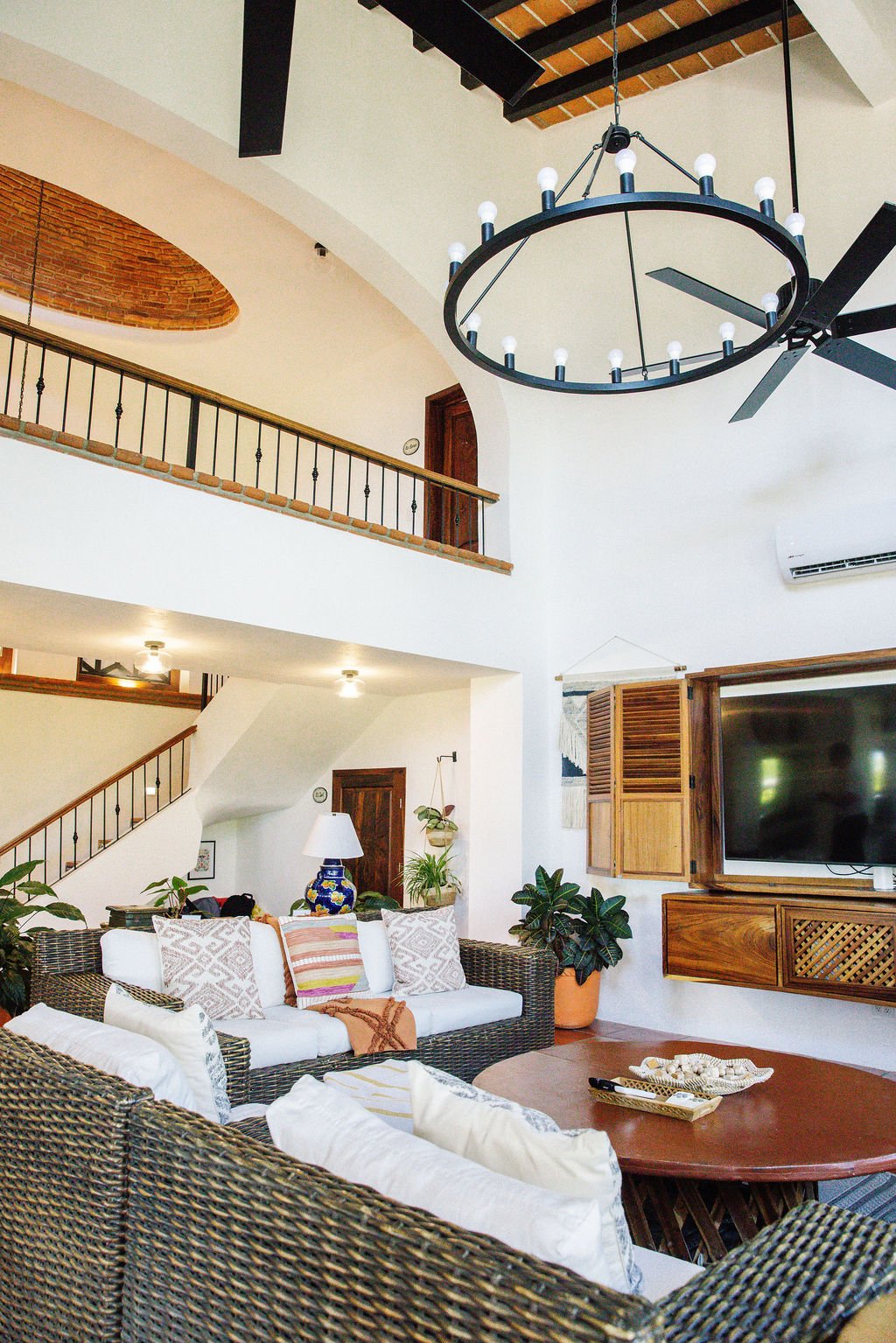 A living room with wicker sofas, white cushions, a wooden coffee table, potted plants, a wall-mounted TV, and a black chandelier with multiple bulbs hanging from the ceiling.