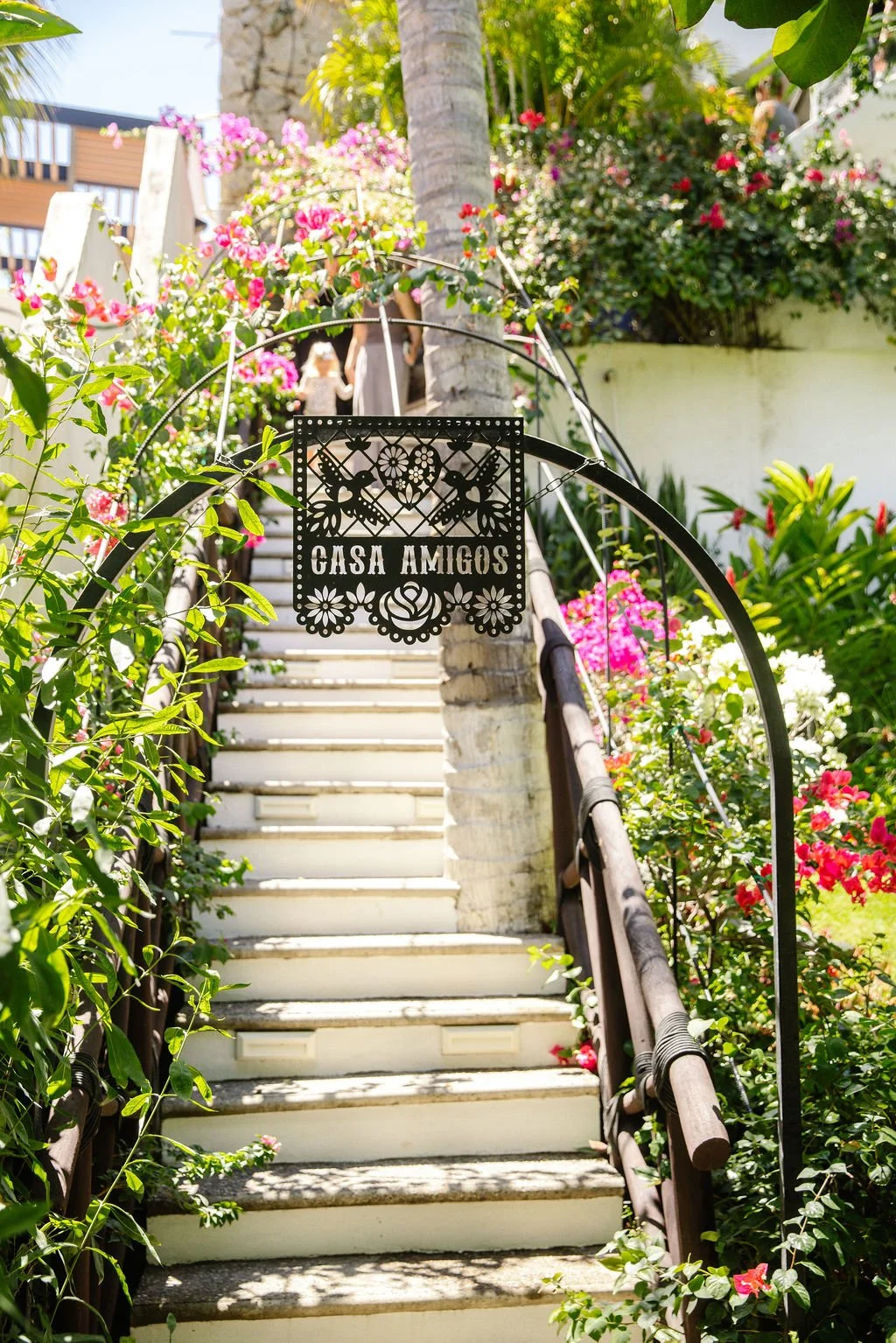 Outdoor staircase surrounded by lush greenery and pink, white, and red flowers leading up to a building with a sign that reads 'Casa Amigos' hanging in the middle of the staircase.