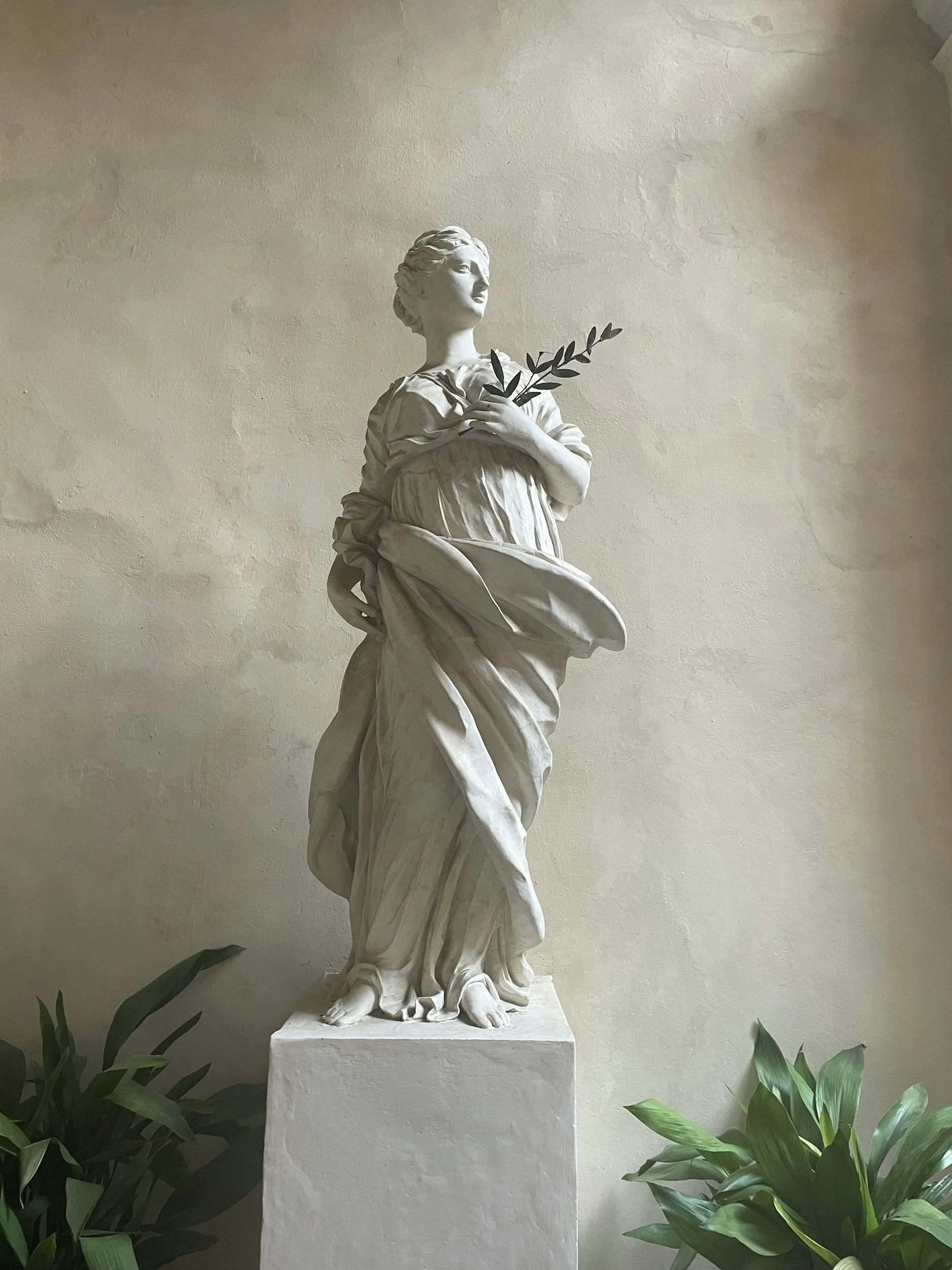 Statue of a woman holding a branch, standing on a pedestal surrounded by green plants.