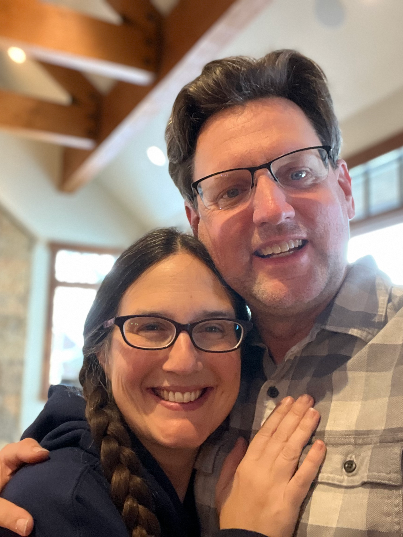 A smiling woman and man wearing glasses are close together, embracing in a cozy indoor setting with warm lighting and wooden beams.