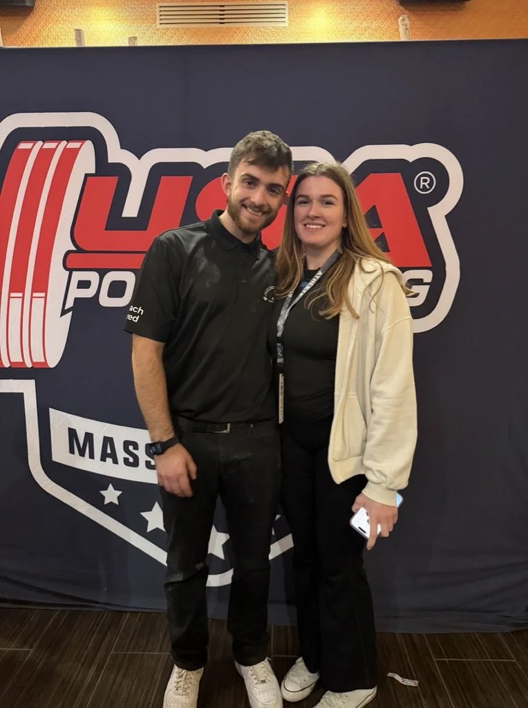 Two young adults, a man and a woman, pose together smiling in front of a large, red and black logo for The American Powerlifting Federation.