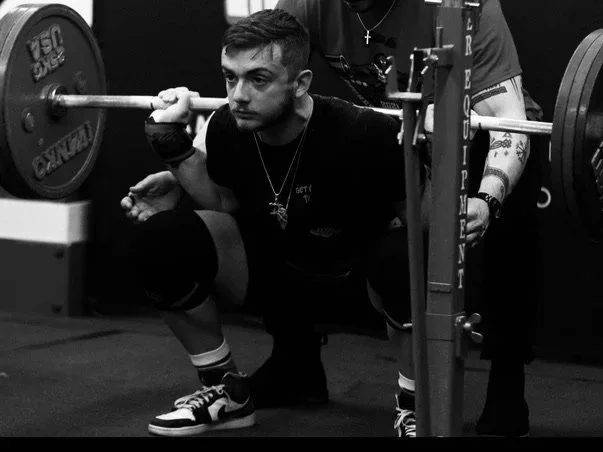 A man lifting a barbell on a squat rack, with a spotter nearby supporting him.