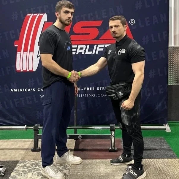 Two men shaking hands in front of a backdrop with a powerlifting logo. One is wearing a black t-shirt and blue pants, the other is dressed in black with a black fanny pack. They are in a gym or competition setting.