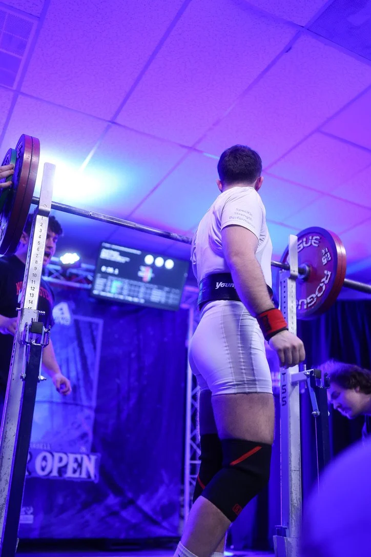 A male powerlifter preparing for a squat lift in a competition setting, wearing a white outfit with a black belt and knee wraps, standing in front of a barbell loaded with weights, with a scoreboard visible in the background.