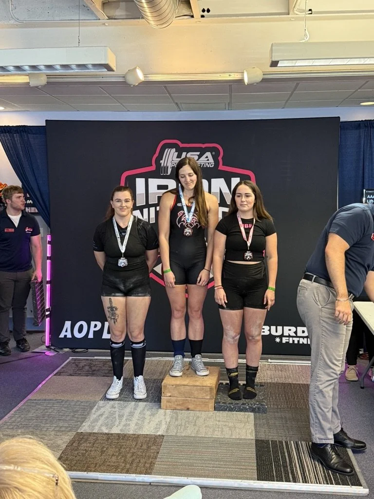 Three women standing on a podium at a powerlifting competition, with medals around their necks. The woman in the center is on the highest platform, indicating first place, while the women on either side are on lower platforms for second and third places. The background features a sign for the USA Powerlifting organization.