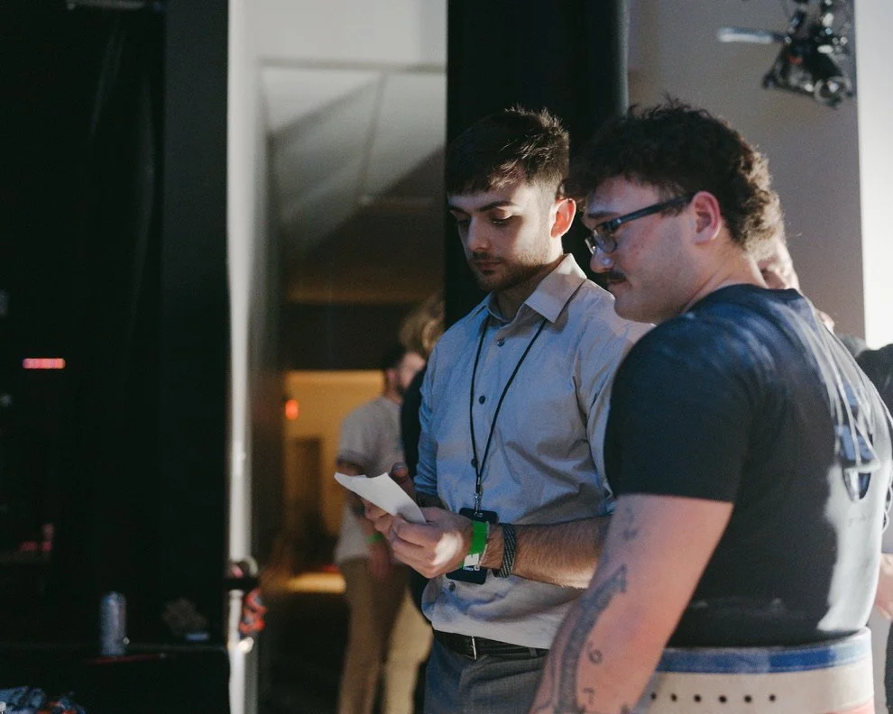 Two men standing close together, looking at a piece of paper or a card in one of their hands, in a dimly lit indoor setting, possibly at an event or a conference.