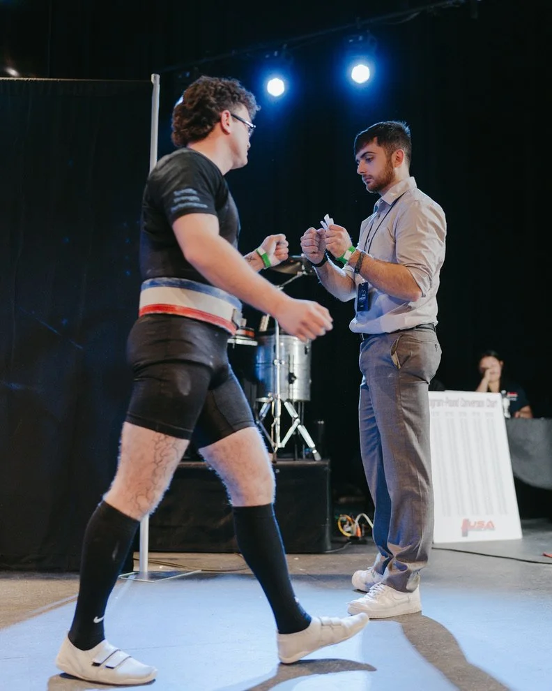 A man in athletic gear, including shorts, socks, and sneakers, is on a stage in a fighting stance, facing another man in a dress shirt and slacks who is holding a small paper. Stage lights are overhead, and there is equipment and a person in the back
