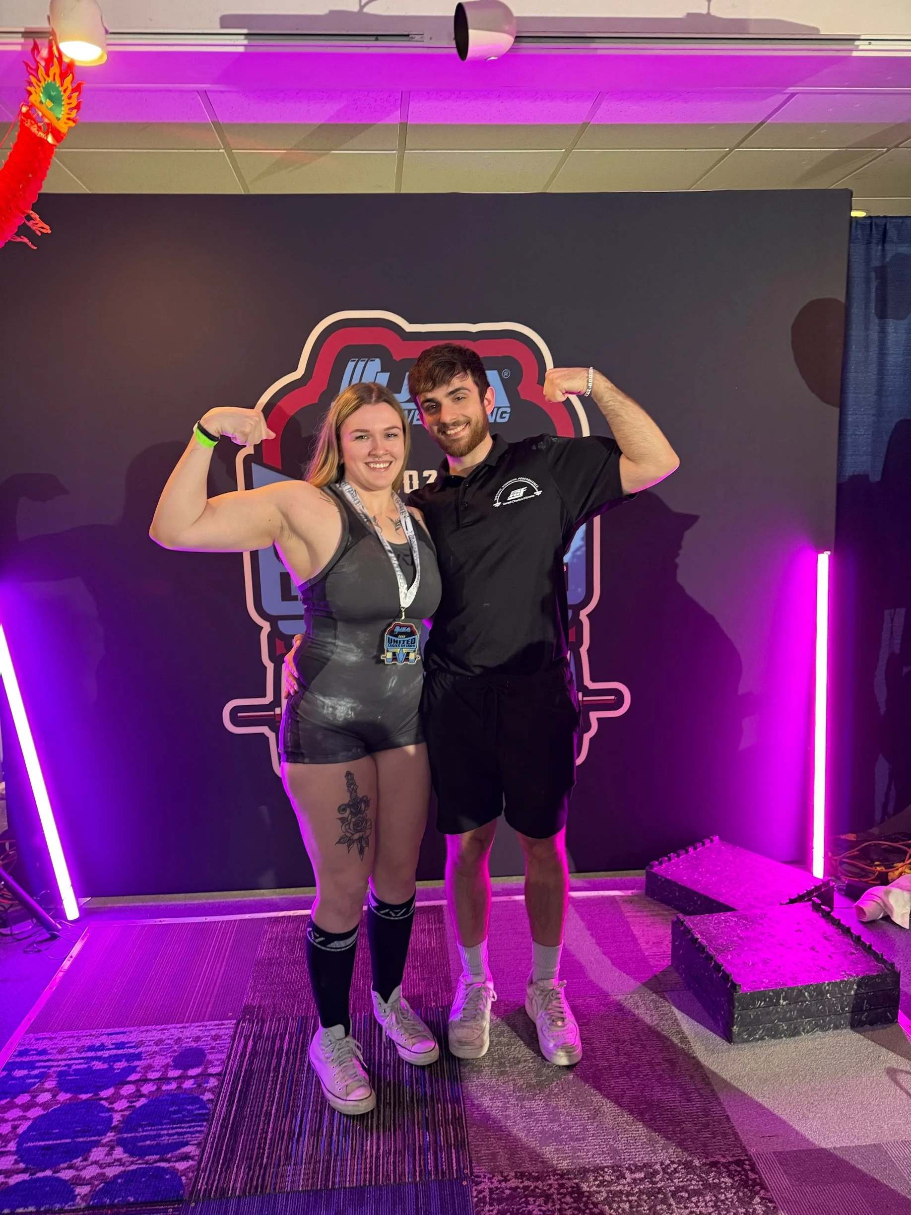 A man and a woman are posing in front of a backdrop at a fitness event, both flexing their arm muscles and smiling. The woman is wearing a sports top and shorts, with tattoos on her left thigh, while the man is dressed in a black shirt and shorts. The background features a logo and purple lighting.