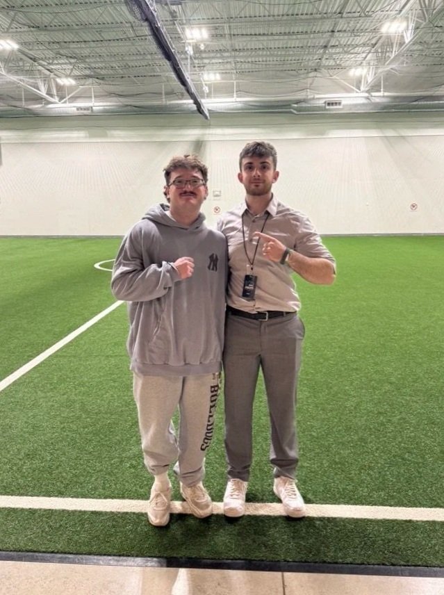 Two men standing on an indoor soccer field, one wearing a gray hoodie and sweatpants, the other in a dress shirt and pants, posing together with their fists raised.
