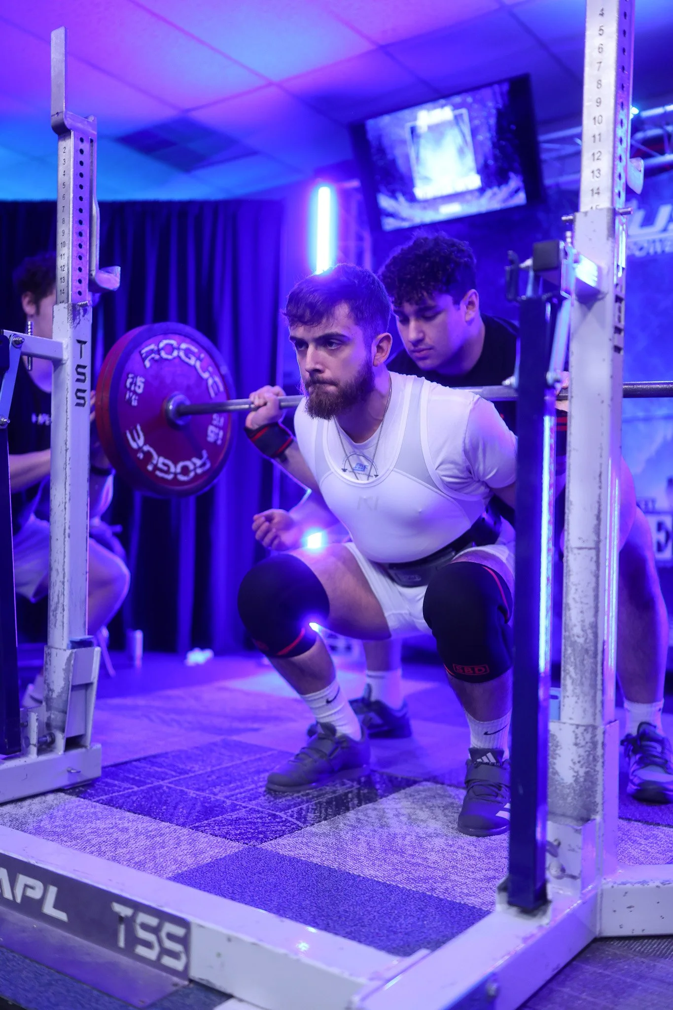A man with a beard and white athletic clothing is squatting and performing a barbell squat, supported by two spotters. The setting is a gym with purple and blue lighting.