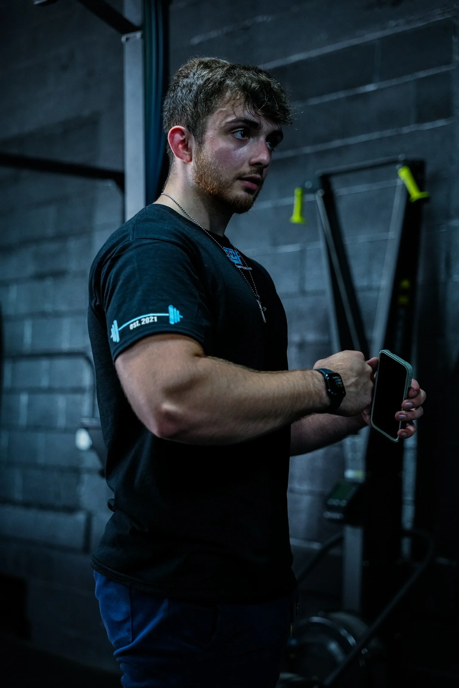 A young man with brown hair and a beard wearing a black T-shirt with a fitness logo, standing in a gym, using a smartphone.