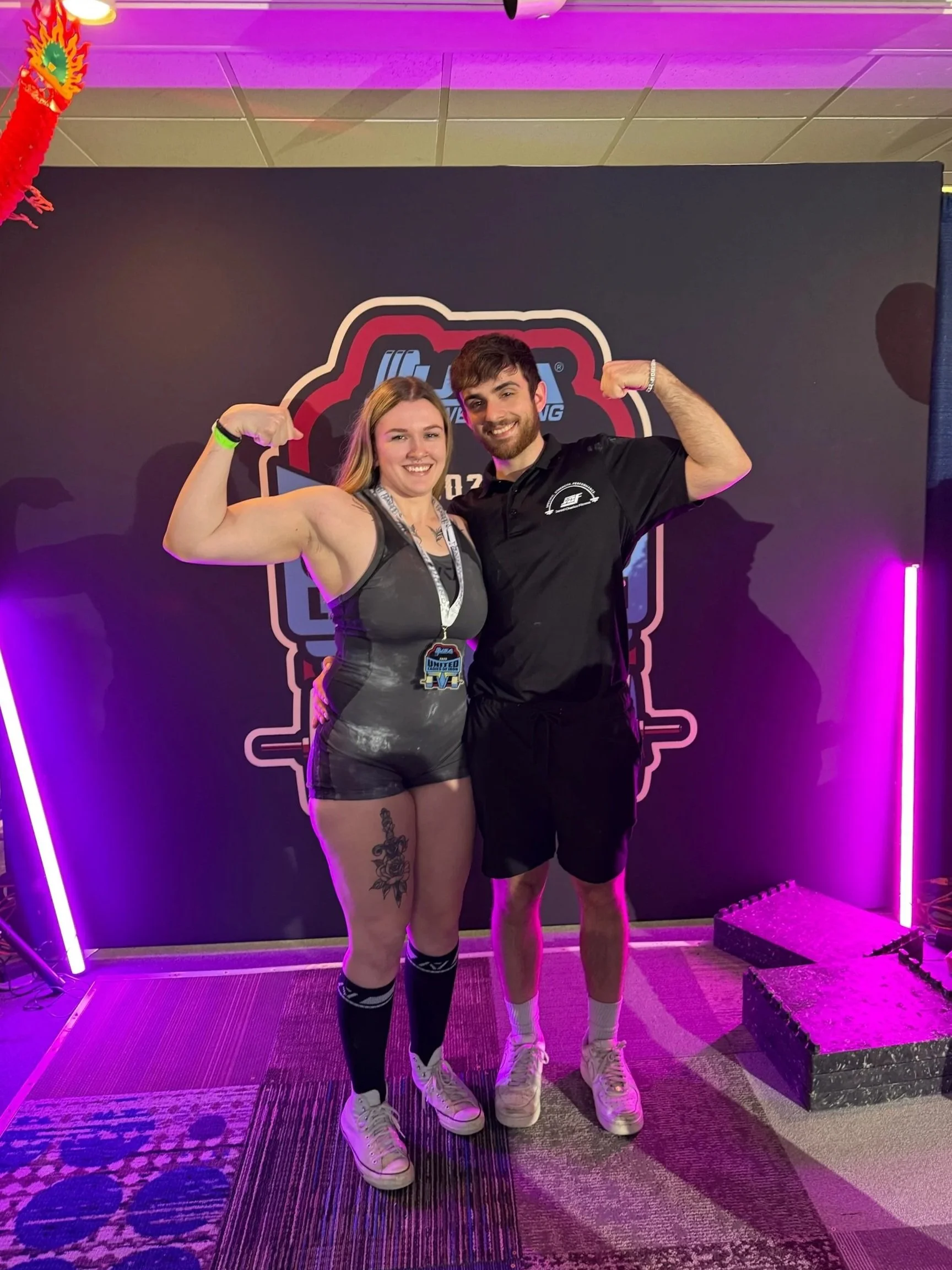 A woman and a man are flexing their biceps and smiling at the camera in front of a backdrop with a logo. The woman has tattoos on her thigh, is wearing a black athletic outfit, and white sneakers. The man is wearing a black polo shirt, black shorts, and white sneakers. They are in a room with purple lighting and decorative neon lights.