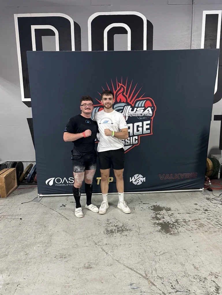 Two men stand side by side in a gym, smiling and flexing their biceps in front of a backdrop with a competition logo that reads 'US Powerlifting Edge Classic'. One man is wearing a black shirt, shorts, and leg compression sleeves, while the other is 