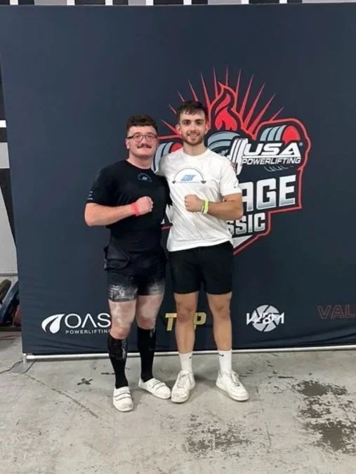 Two young men standing in front of a USA Powerlifting stage backdrop, both smiling and posing with their fists raised. One is wearing black shorts, a black T-shirt, and knee-high compression sleeves. The other is dressed in a white T-shirt, black shorts, and white sneakers.