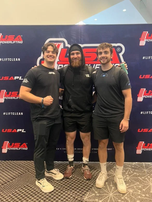 Three young men standing in front of a USA Powerlifting backdrop at a lifting event, smiling and posing for the photo.