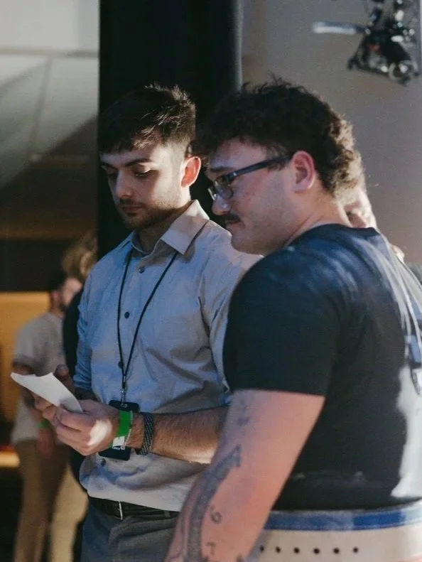 Two men standing close together indoors, one is holding a paper, the other has tattoos on his arm and a mustache, with blurred people in the background.