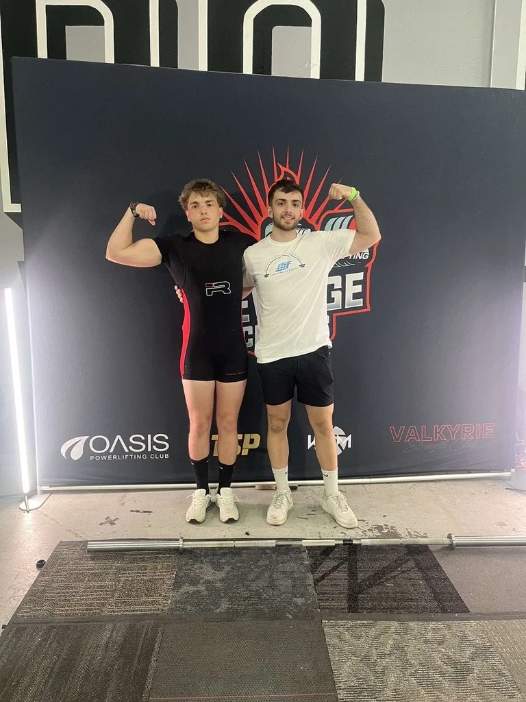 Two young men posing in front of a banner at a powerlifting event, with one wearing a black powerlifting suit and the other in casual sportswear, both flexing their arm muscles.