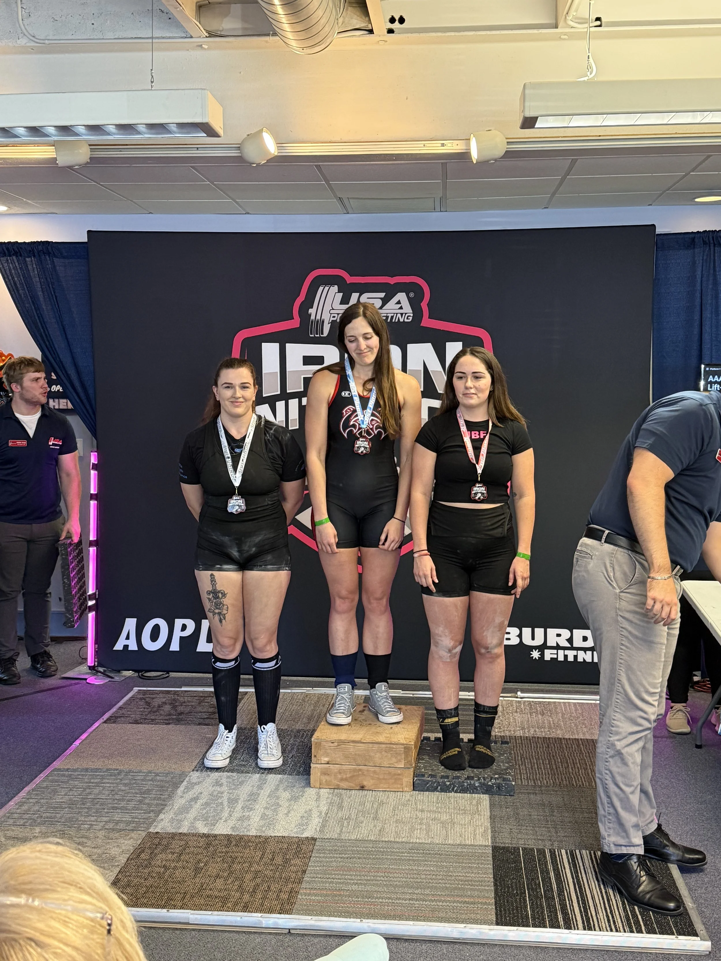 Three women standing on a winners' podium, medaled in a bodybuilding or powerlifting competition, with a venue background featuring a USA Powerlifting sign.