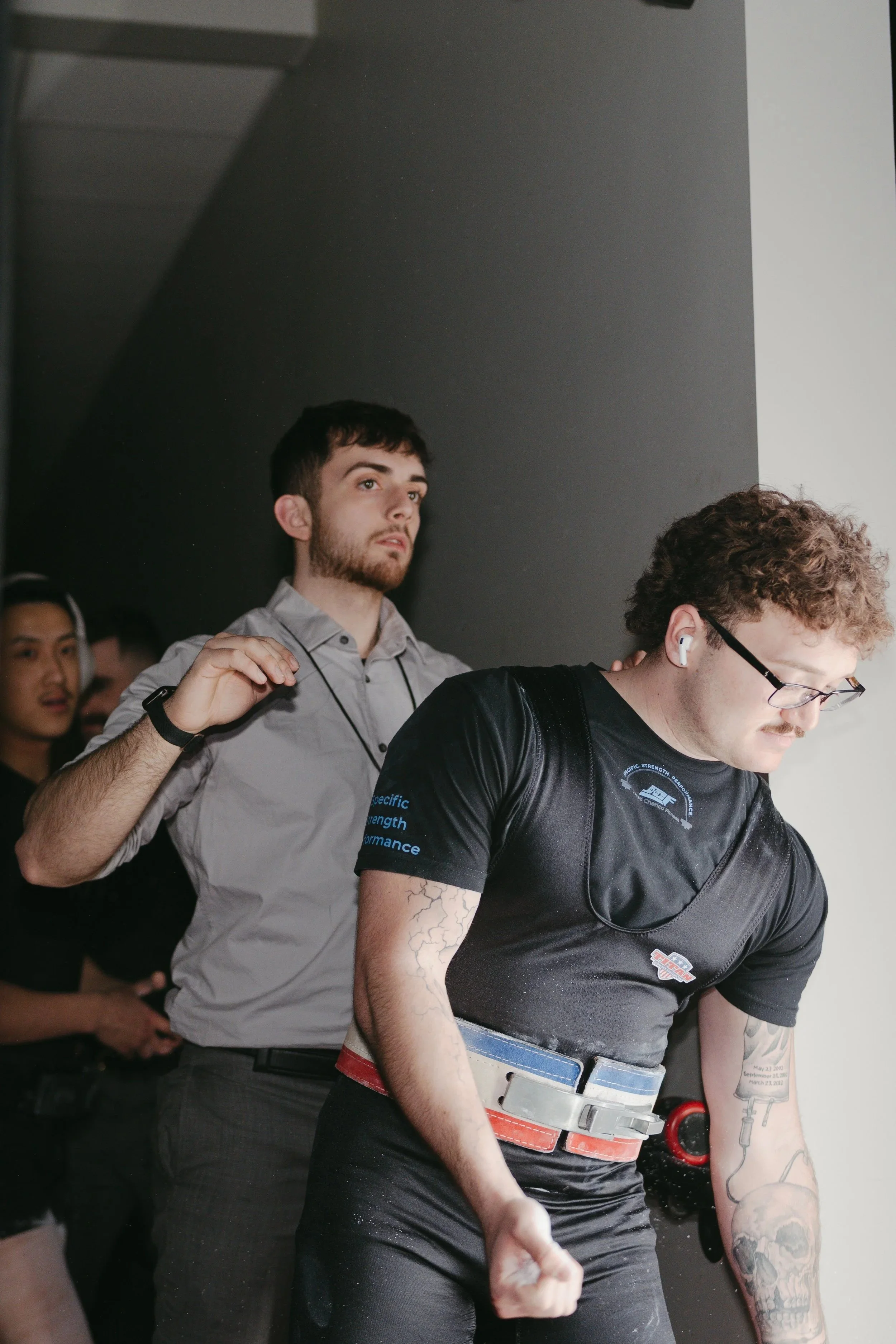 A group of young people, mostly men, standing close together indoors. The focus is on a young man with dark hair and a beard, wearing a light gray shirt, and a young man with curly hair and glasses in a black shirt with a graphic design. The young man with glasses is looking down, and the others are looking in various directions.