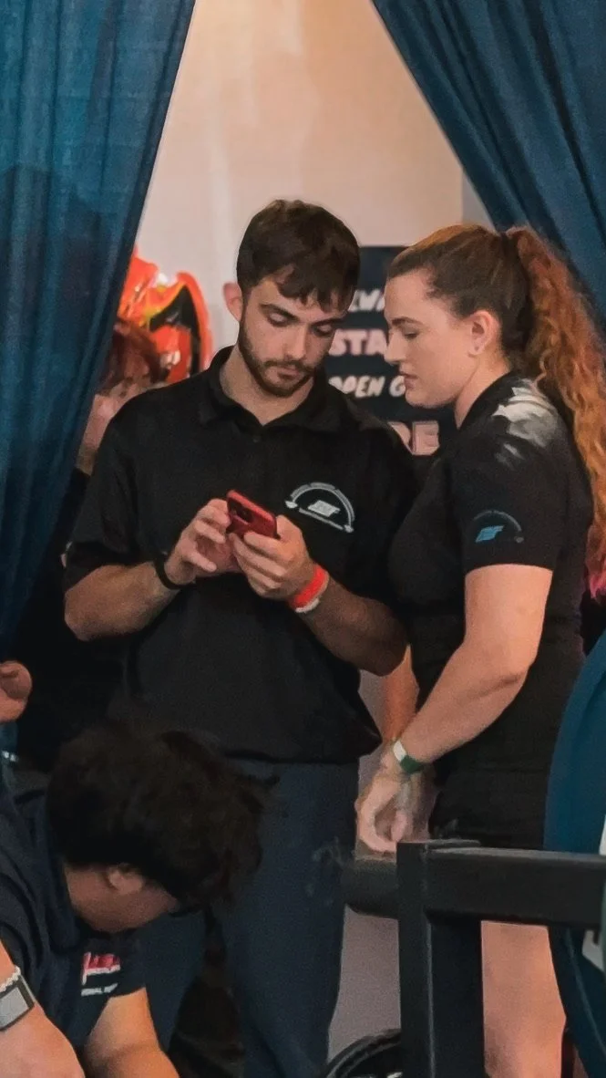 Two people, a man and a woman, are standing close together behind a blue curtain, looking at a phone. The man has dark hair and a beard, and the woman has long curly hair tied back. They appear to be at an event or exhibition, wearing black shirts wi