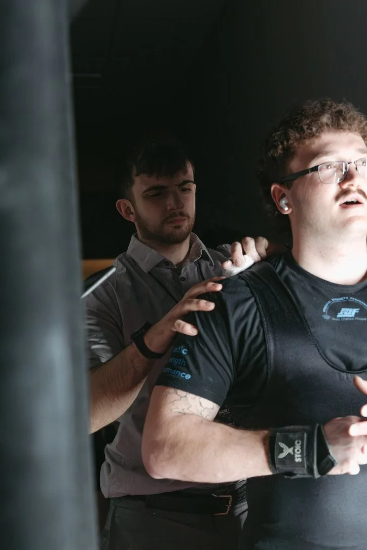 A man assisting a person with earbuds and glasses during a workout.