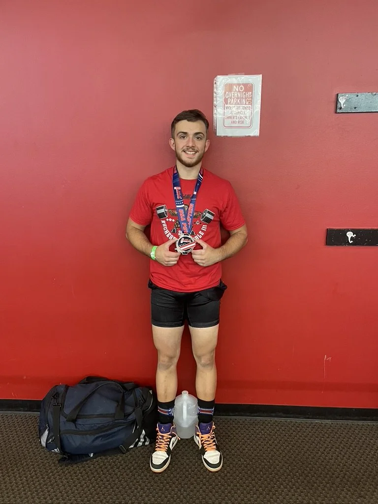 Young man wearing athletic shorts, sneakers, and a medal, standing against a red wall with a backpack at his feet and a water bottle between his legs, smiling at the camera.