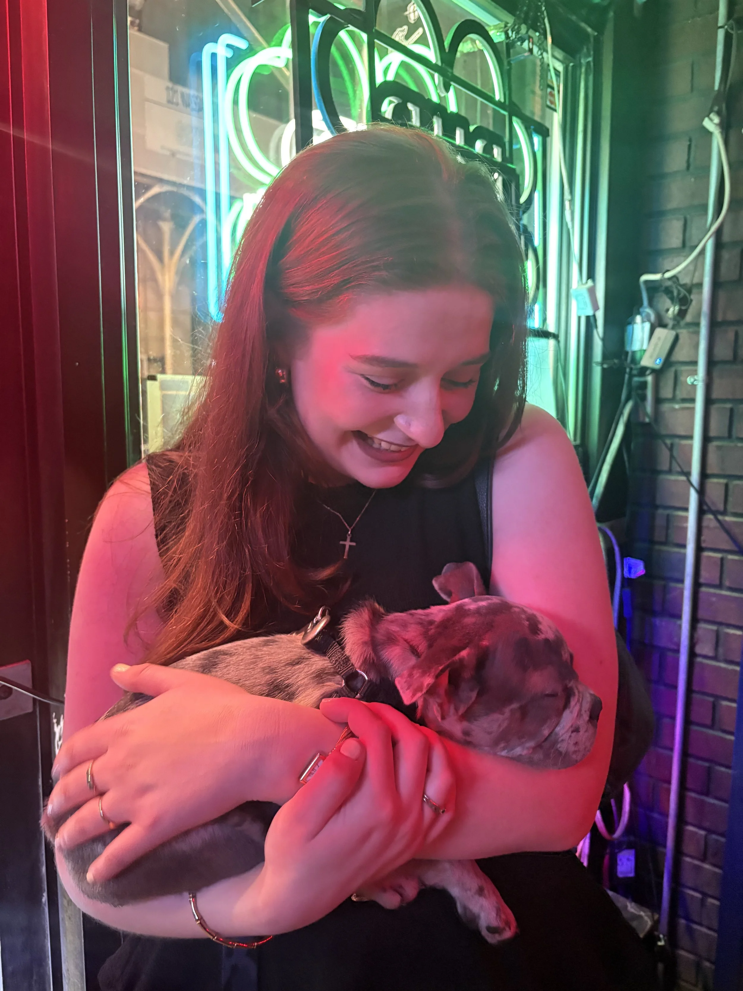 A woman with long reddish-brown hair, wearing a cross necklace and a red bracelet, is smiling while holding a small, sleeping puppy in her arms. The background shows neon signs and a brick wall illuminated by colorful lights.