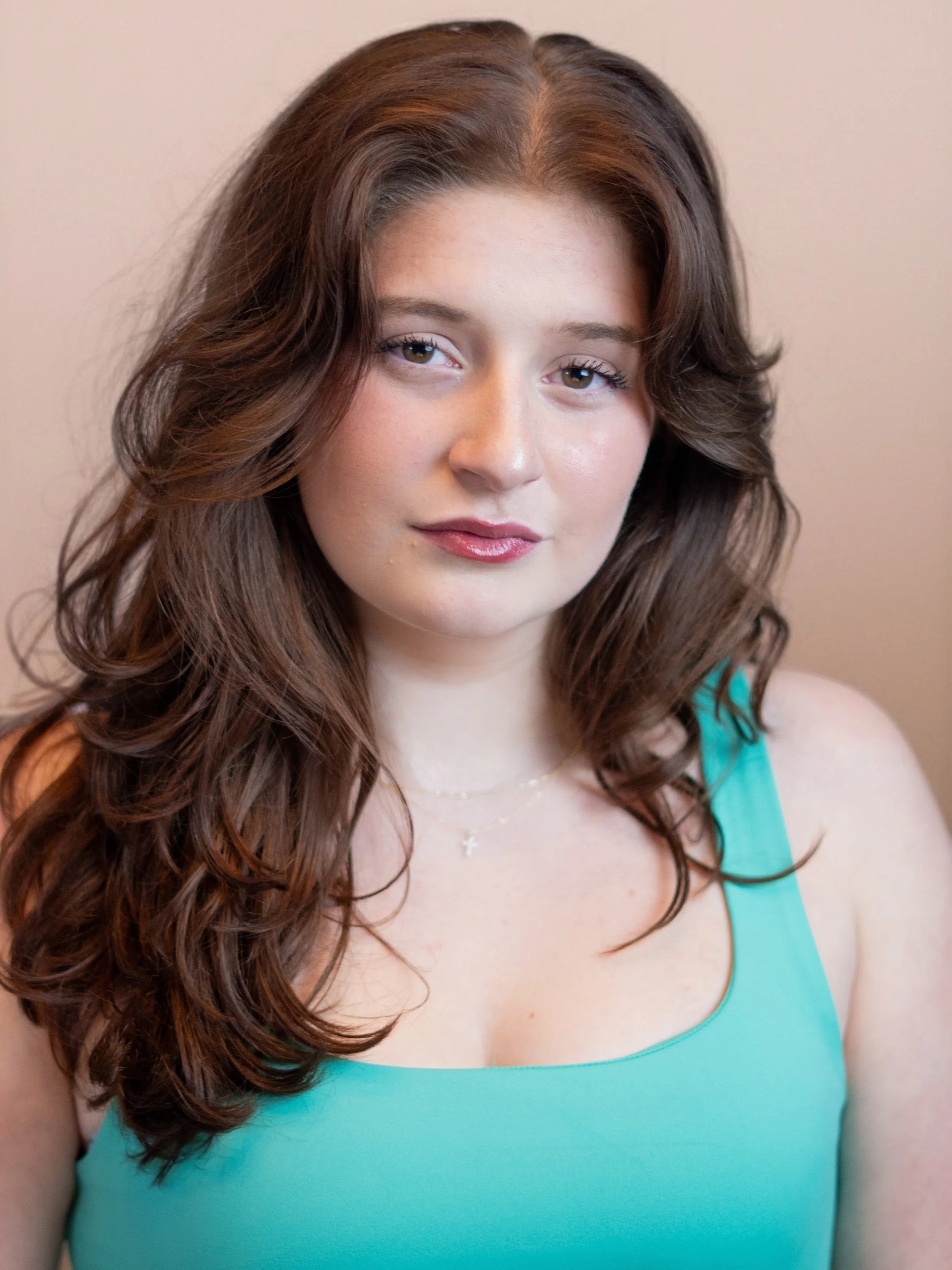 Close-up of a young woman with long, wavy brown hair wearing a turquoise sleeveless top and a delicate necklace with a small cross pendant, standing against a plain background.