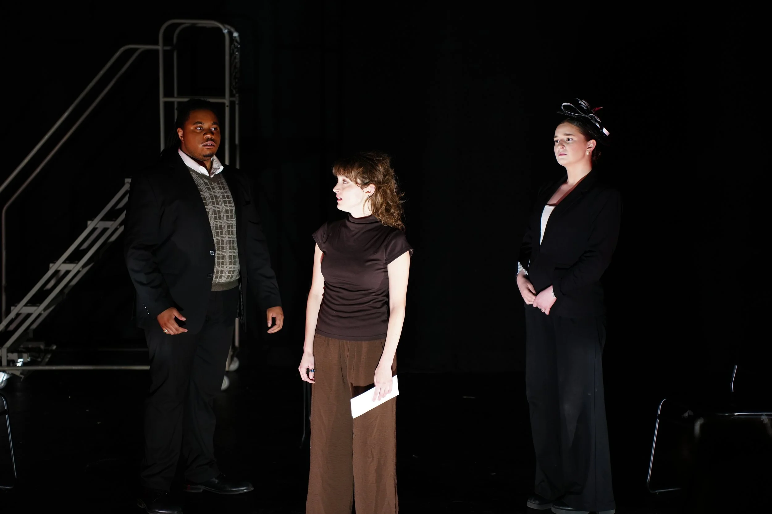 Three people standing in a dark environment with a metal staircase in the background. They are dressed formally and looking in different directions. The woman on the right is wearing a black hat with ribbons, the woman in the center is holding papers, and the man on the left is wearing a black jacket over a brown plaid vest.